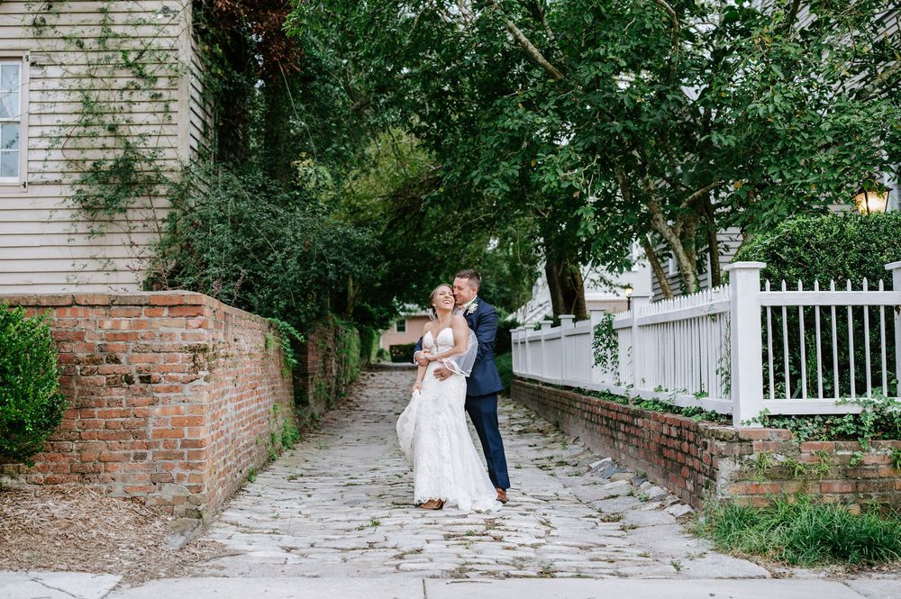 A couple shares a romantic moment on a brick-lined garden path with white picket fence and greenery.