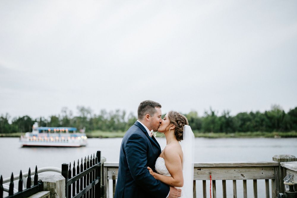 A couple shares a romantic moment on a waterfront deck with a boat and trees visible in the background.