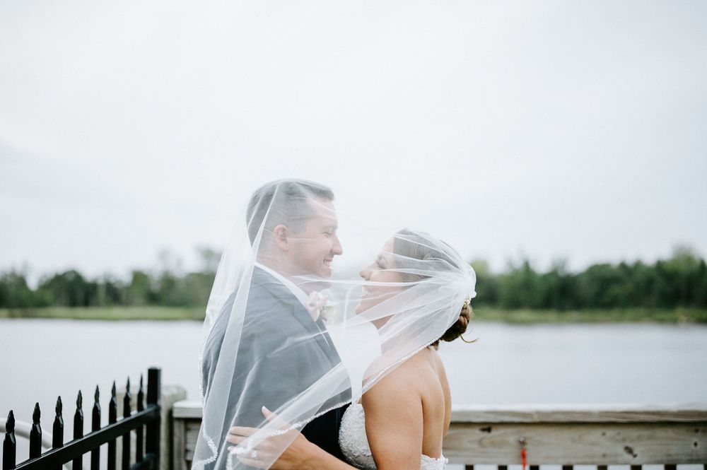 A romantic moment between newlyweds sharing a kiss under a flowing veil by a waterfront with trees in the background.