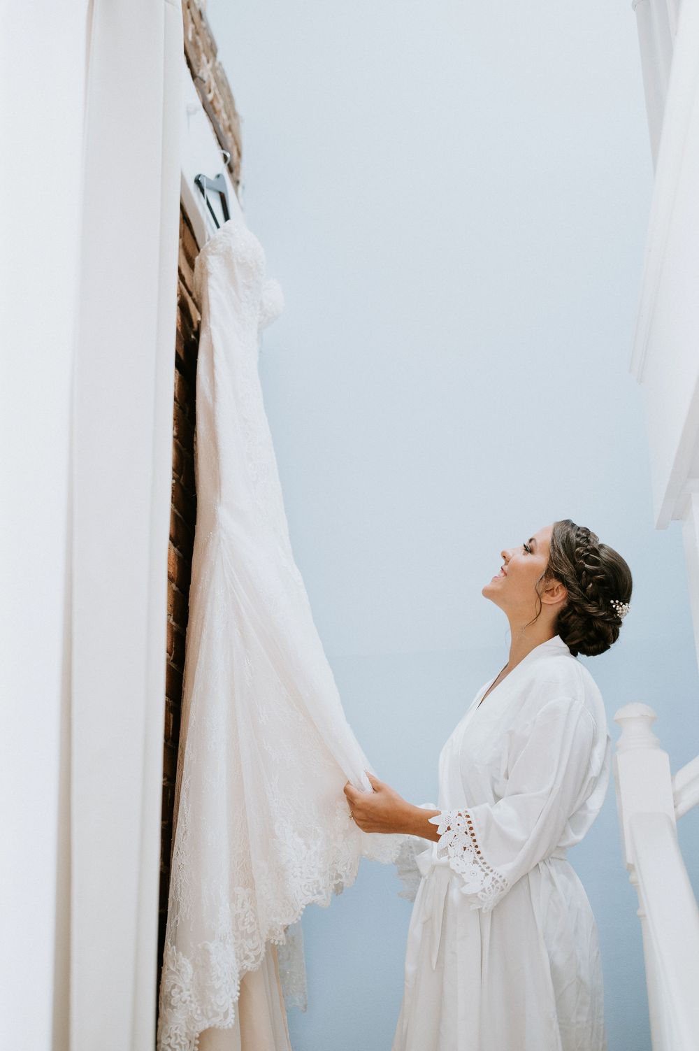 A bride in a white robe admires her wedding dress hanging against a light blue wall during wedding preparations.