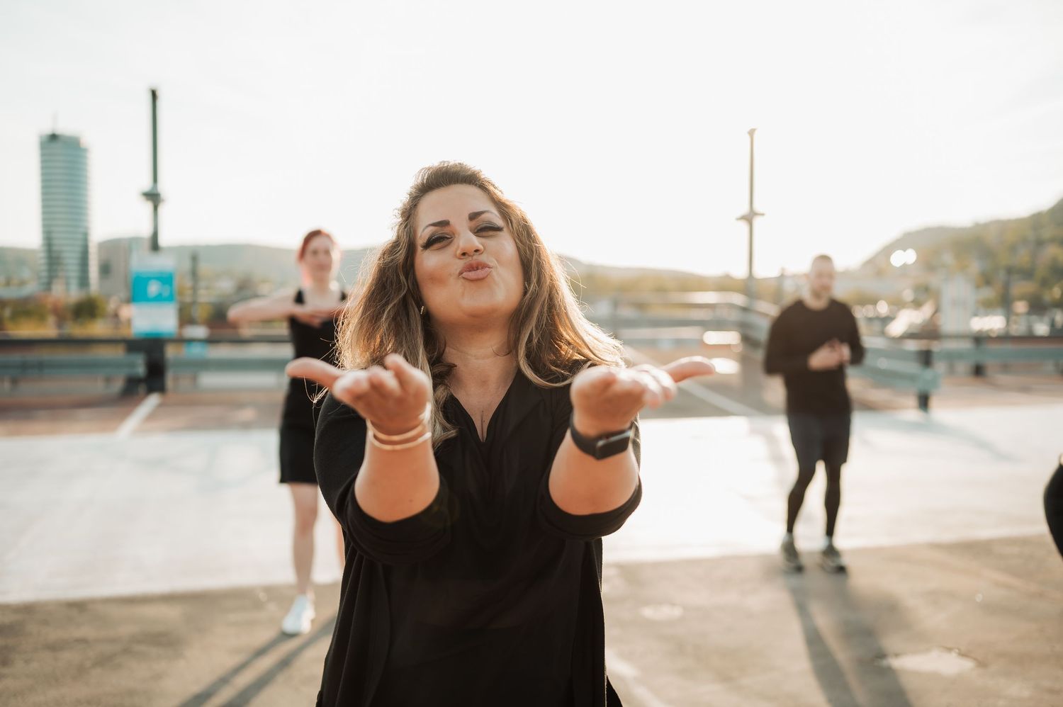 Person in black shirt poses expressively outdoors during golden hour with blurred figures in background.
