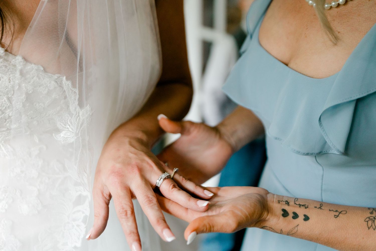 Bride and woman in blue dress exchanging wedding rings, close-up of hands with tattoos and lace gown.