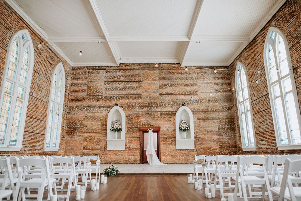 A rustic wedding venue with exposed brick walls, arched windows, and white chairs arranged in rows for a ceremony.