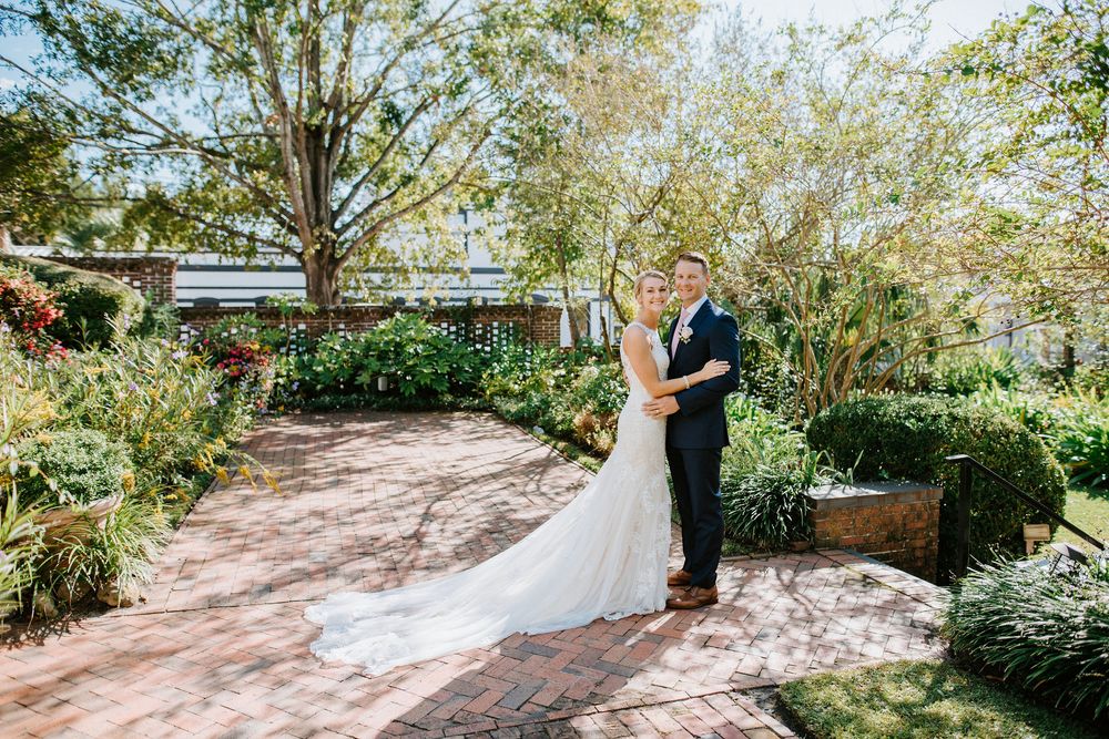 A romantic garden wedding ceremony takes place on a sunlit brick path surrounded by lush greenery and trees.