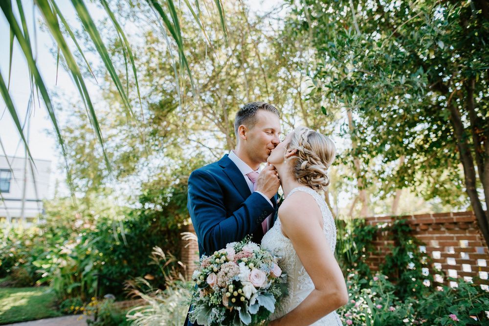 A couple shares a romantic kiss in a lush garden setting with natural sunlight filtering through the trees.