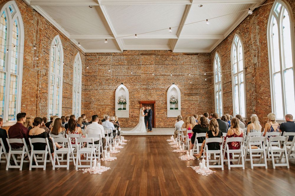 Wedding ceremony in a historic brick chapel with arched windows, wooden floors and white folding chairs for guests.