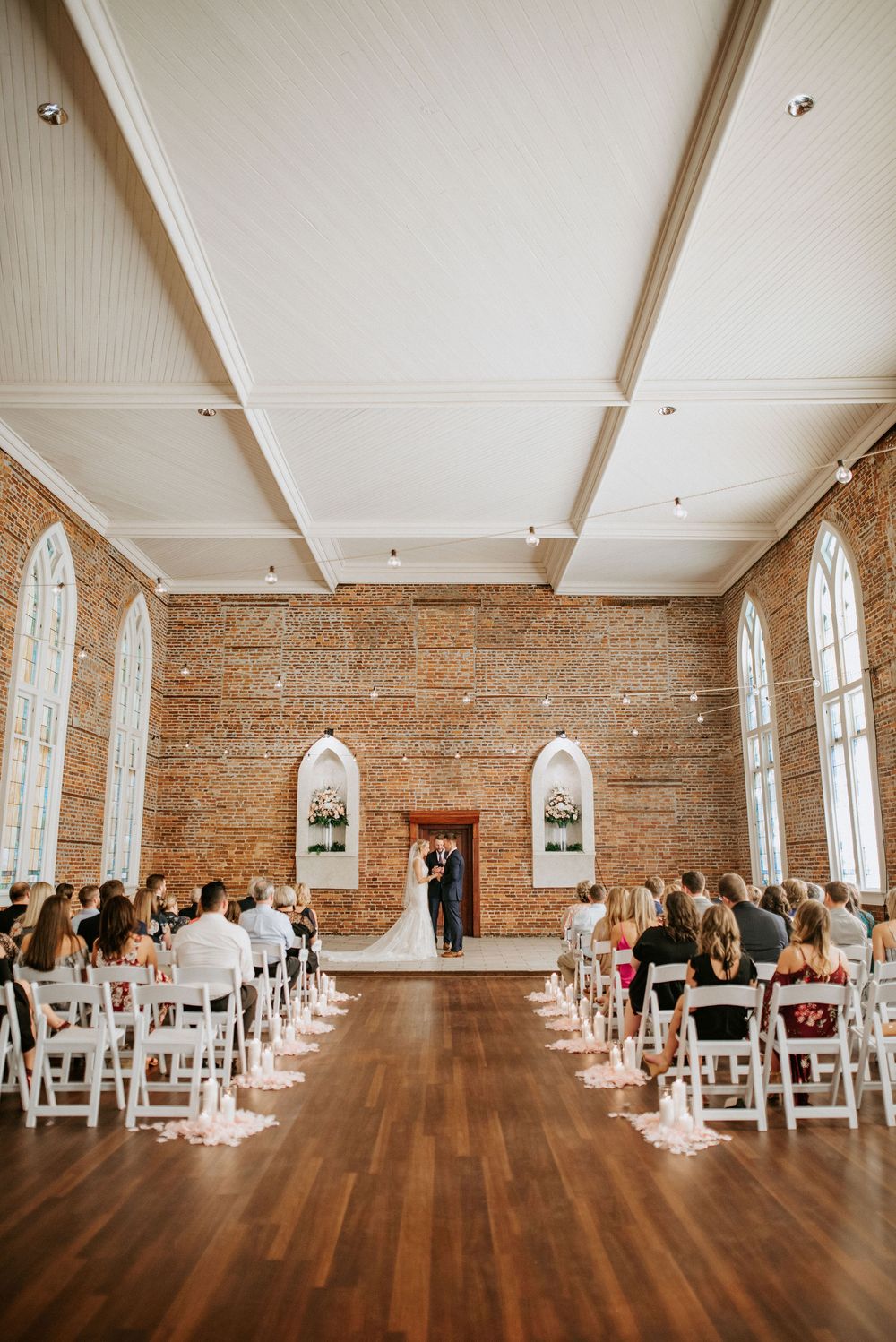 Wedding ceremony setup in a rustic brick venue with white chairs, exposed beams, and natural light streaming through windows.