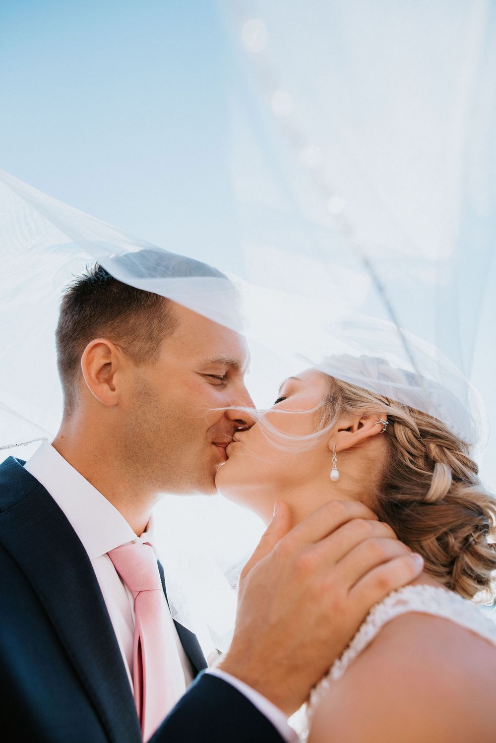 Romantic wedding couple sharing an intimate kiss against a bright blue sky background.
