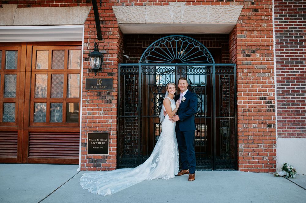 A couple in wedding attire embraces in front of a rustic brick building with ornate wooden doors and windows.