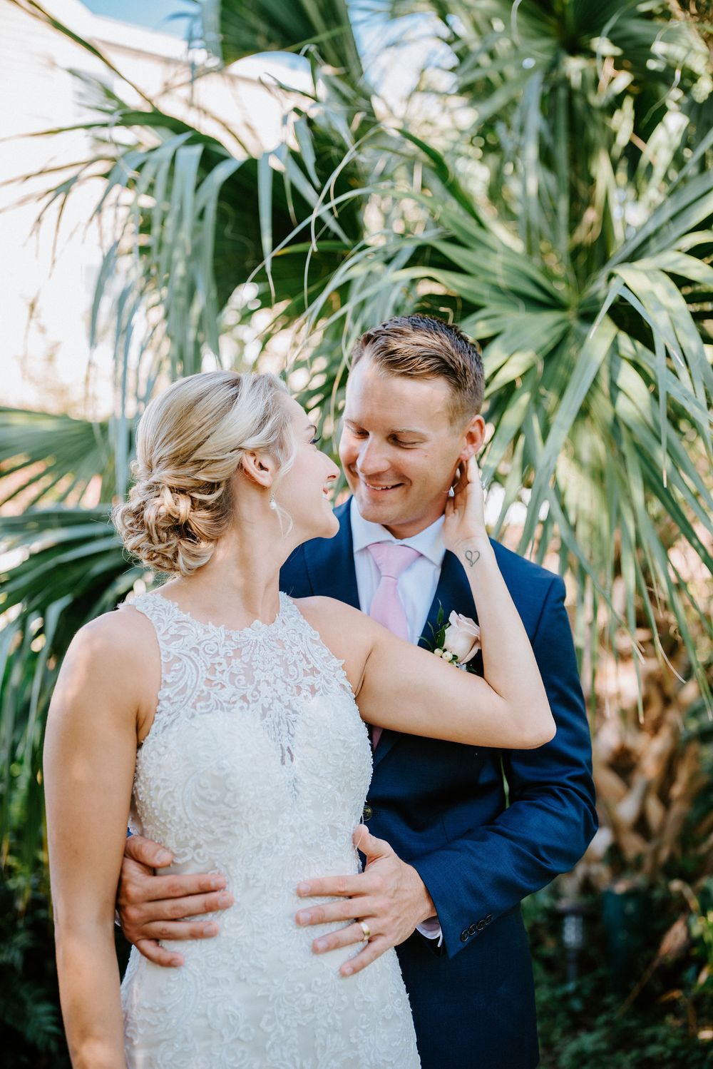 A romantic wedding couple shares tender moments in a lush tropical garden setting with palm trees in the background.