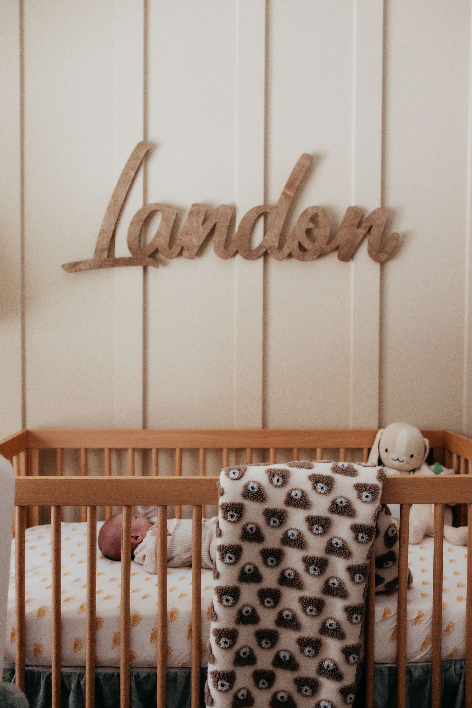 A rustic wooden wall with a decorative name sign above three wooden cribs with matching patterned bedding.