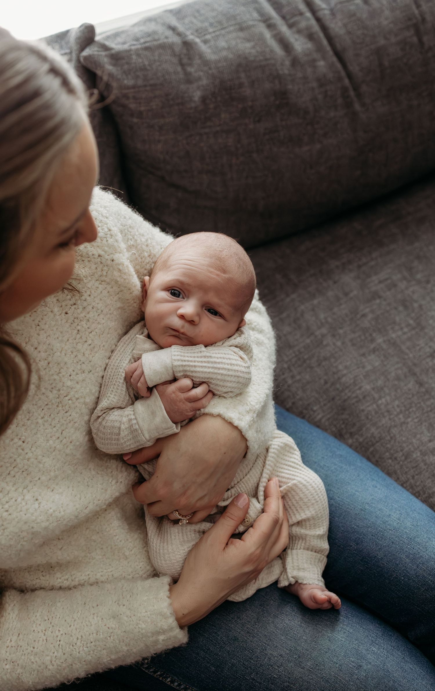 A tender moment on a gray couch as a baby in a cream sweater is held close in loving arms.