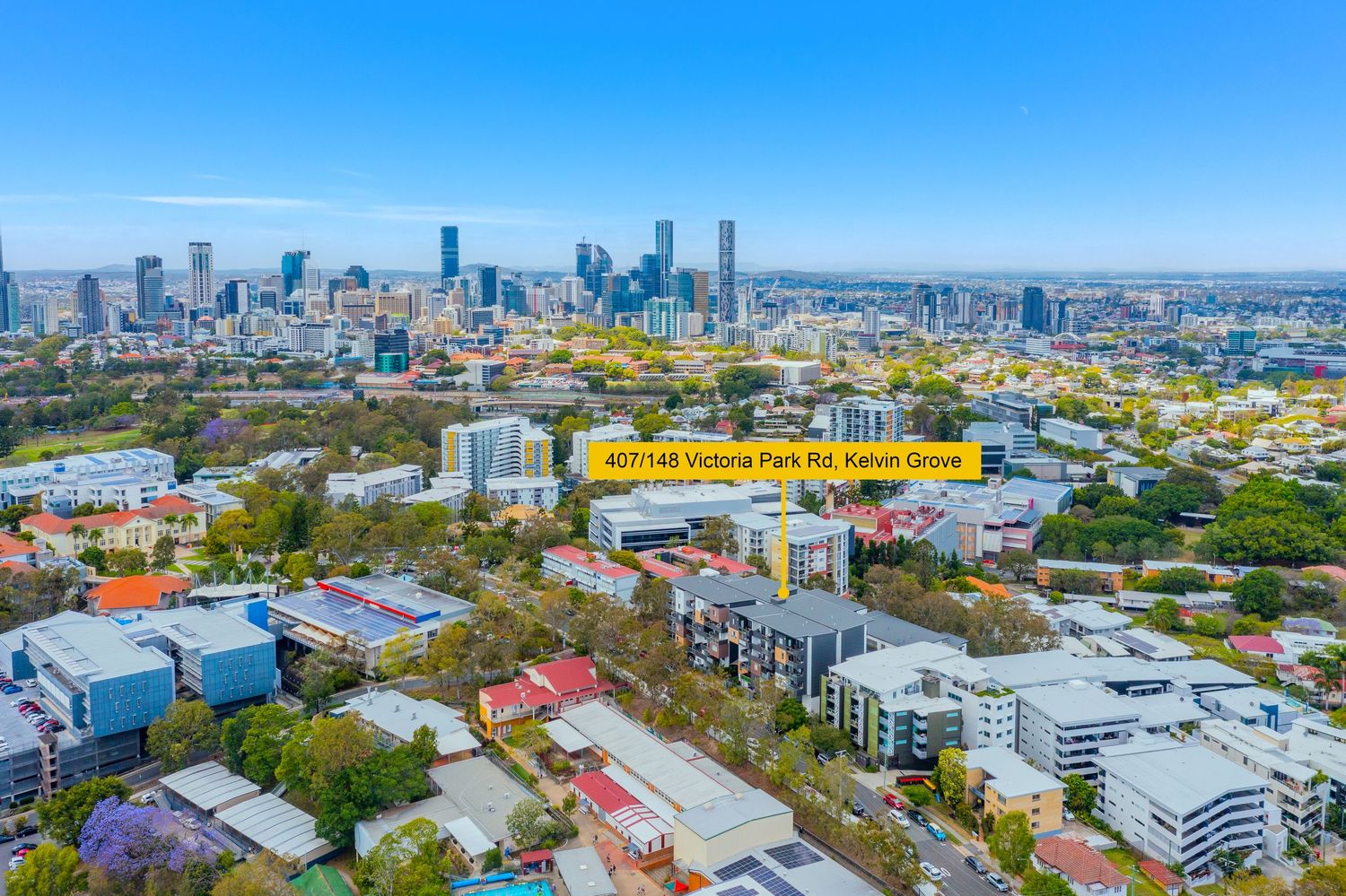 Aerial view of Victoria Park Rd Kelvin Grove with Brisbane CBD skyline backdrop and jacaranda trees.