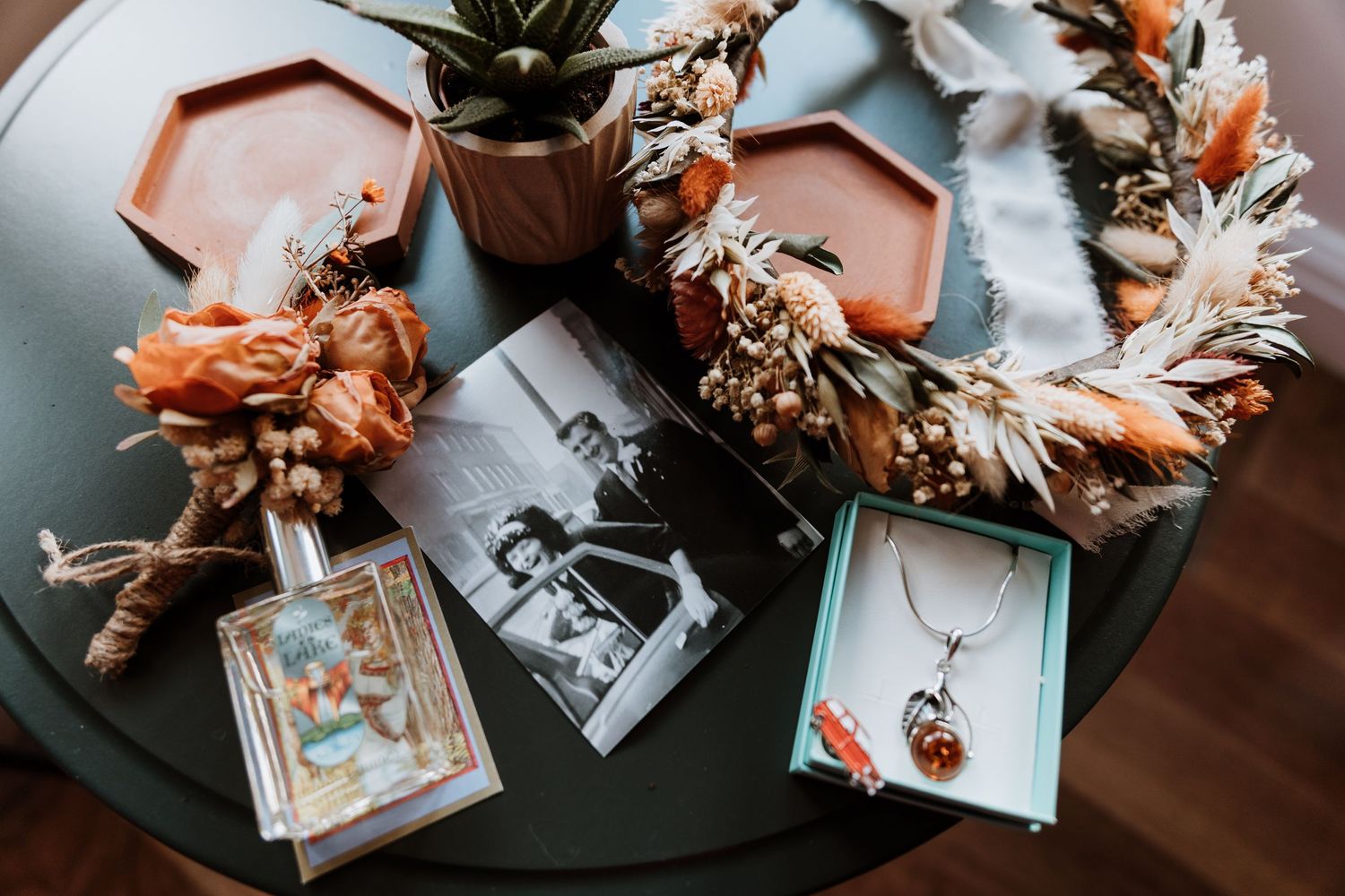 Flatlay of dried flower wreath, vintage photographs, and jewelry on dark green table surface.