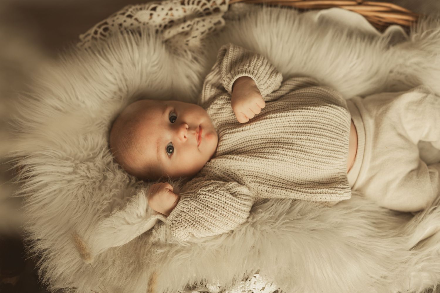 Baby wearing cozy white knit sweater lying on soft furry blanket in warm natural lighting.