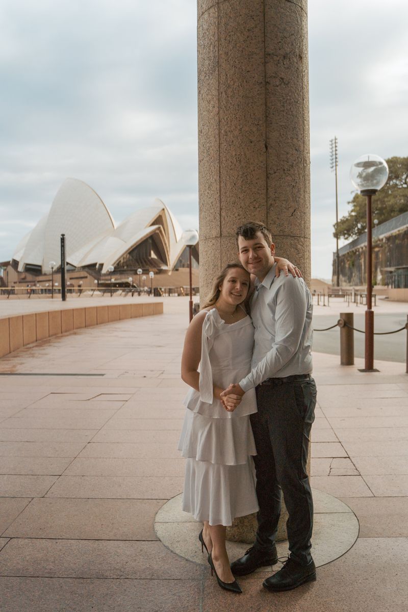 A couple shares a romantic moment under concrete columns with cloudy skies in front of the opera house in sydney their engagement phoshoot