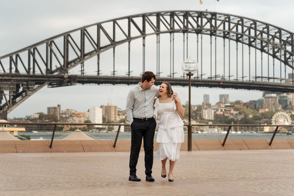 an engaged couple walks hand in hand along Sydney Harbour with iconic bridge and cityscape in background during a photoshoot