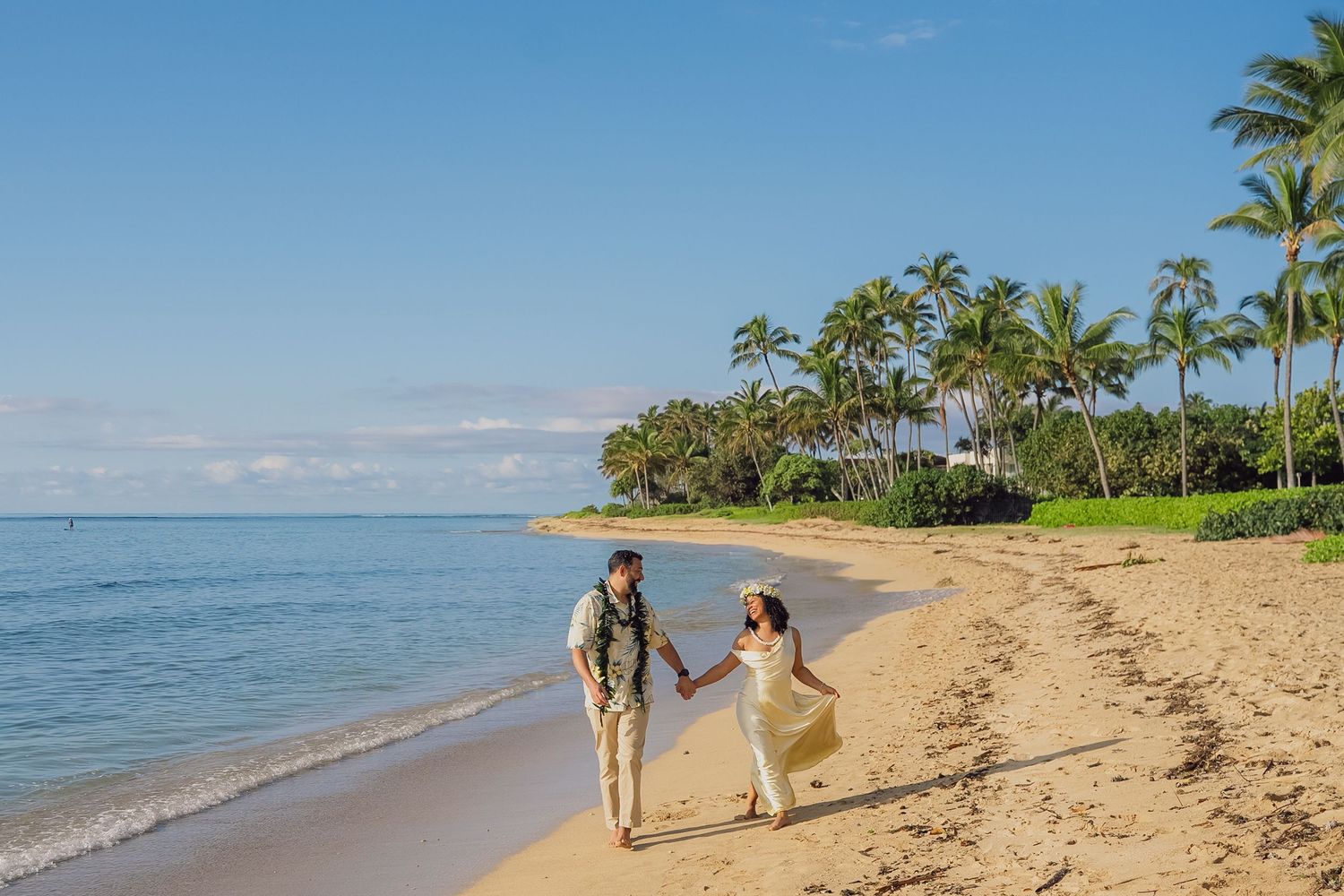 Couple walking hand in hand on a tropical beach with palm trees and blue ocean waters in Hawaii.