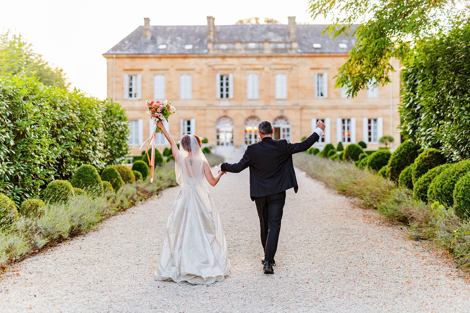 A couple walks hand in hand along a scenic gravel path leading to an elegant French chateau on a sunny day.