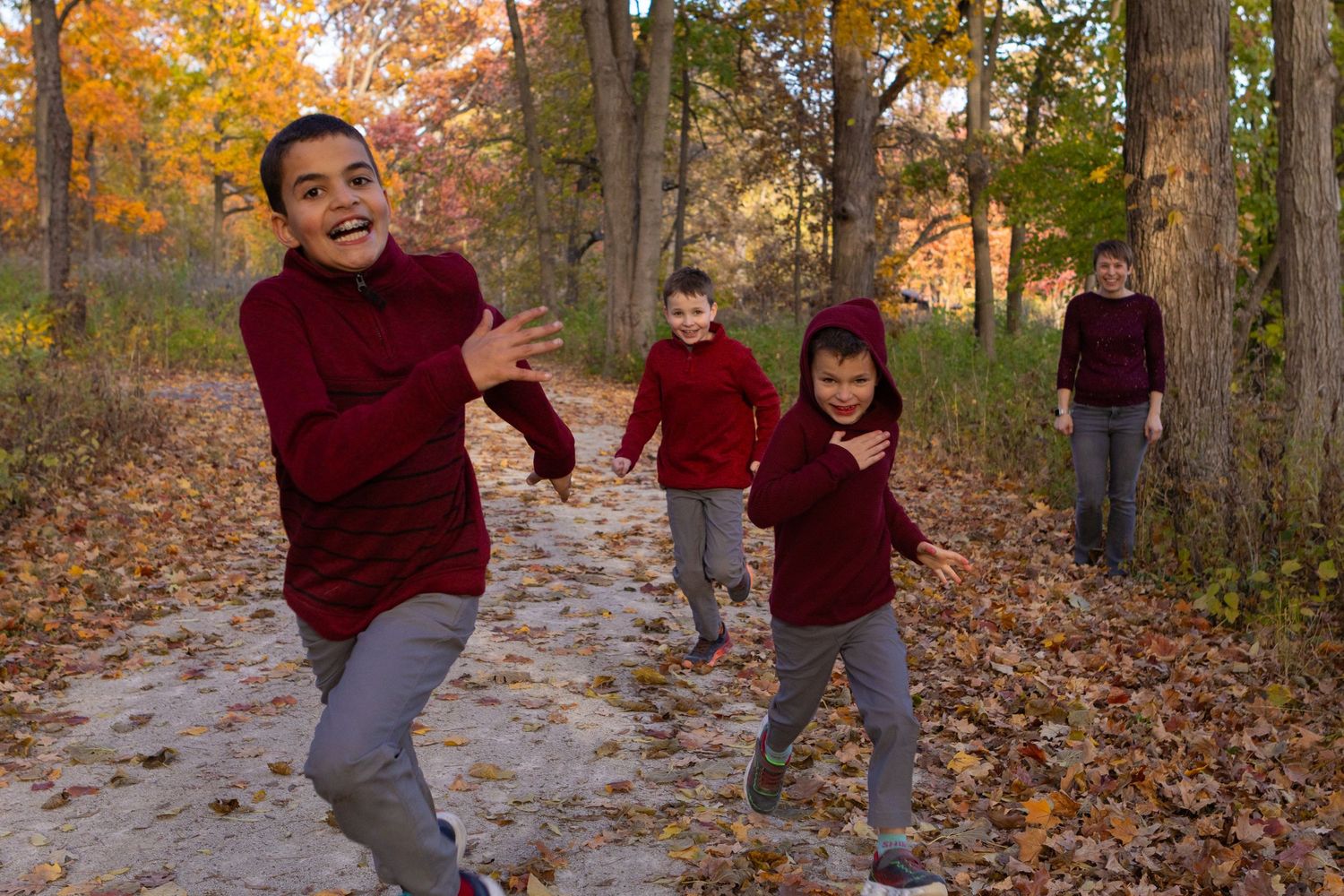 Children in matching burgundy sweaters run and play on a leaf-covered path through autumn woods at sunset.