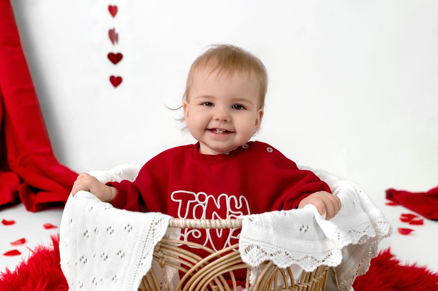 Baby in red and white outfit lying on white blanket with scattered red heart decorations for Valentine's Day themed photo.