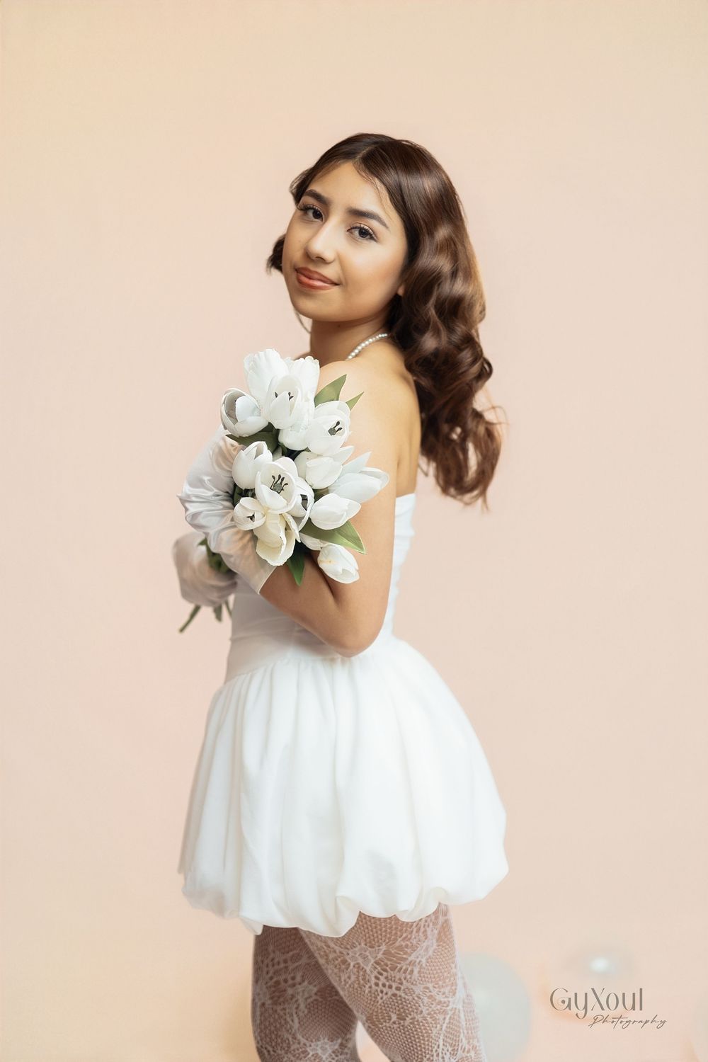 A young girl in a white dress poses gracefully with white flowers against a beige background.
