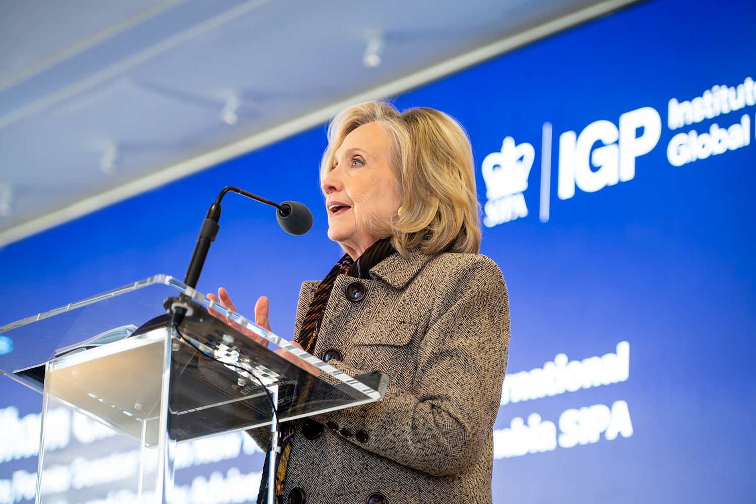 A speaker stands at a podium giving a presentation in front of a blue backdrop with white text.