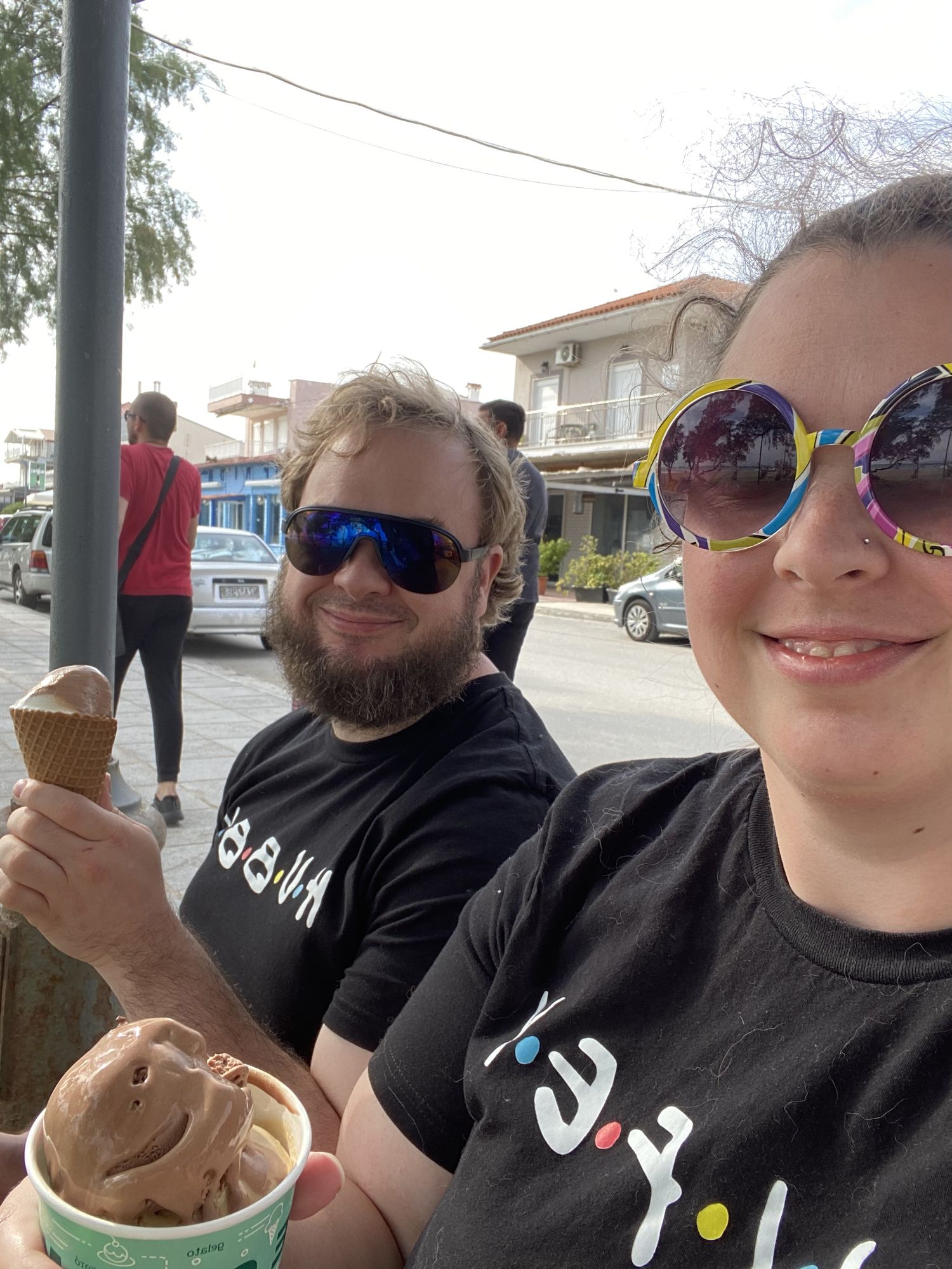 Two people enjoying ice cream cones on a sunny day with cars and buildings visible in the background.