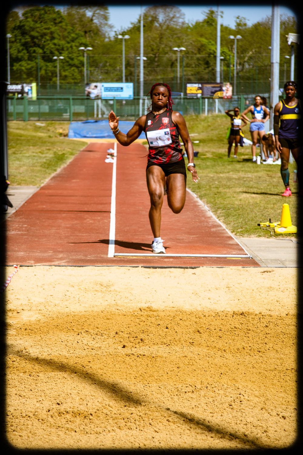 Athletes practice long jump on an outdoor track and field facility with sand pit landing area.