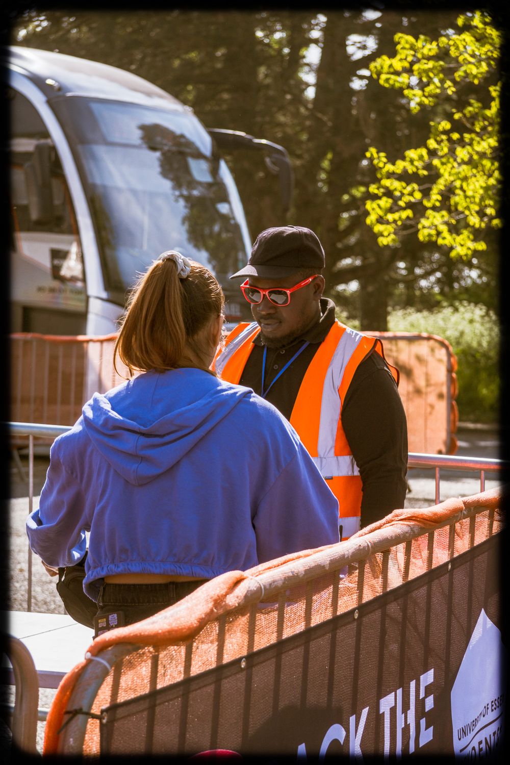 Two people in blue and orange safety vests sit together on a brick wall during a sunny day.