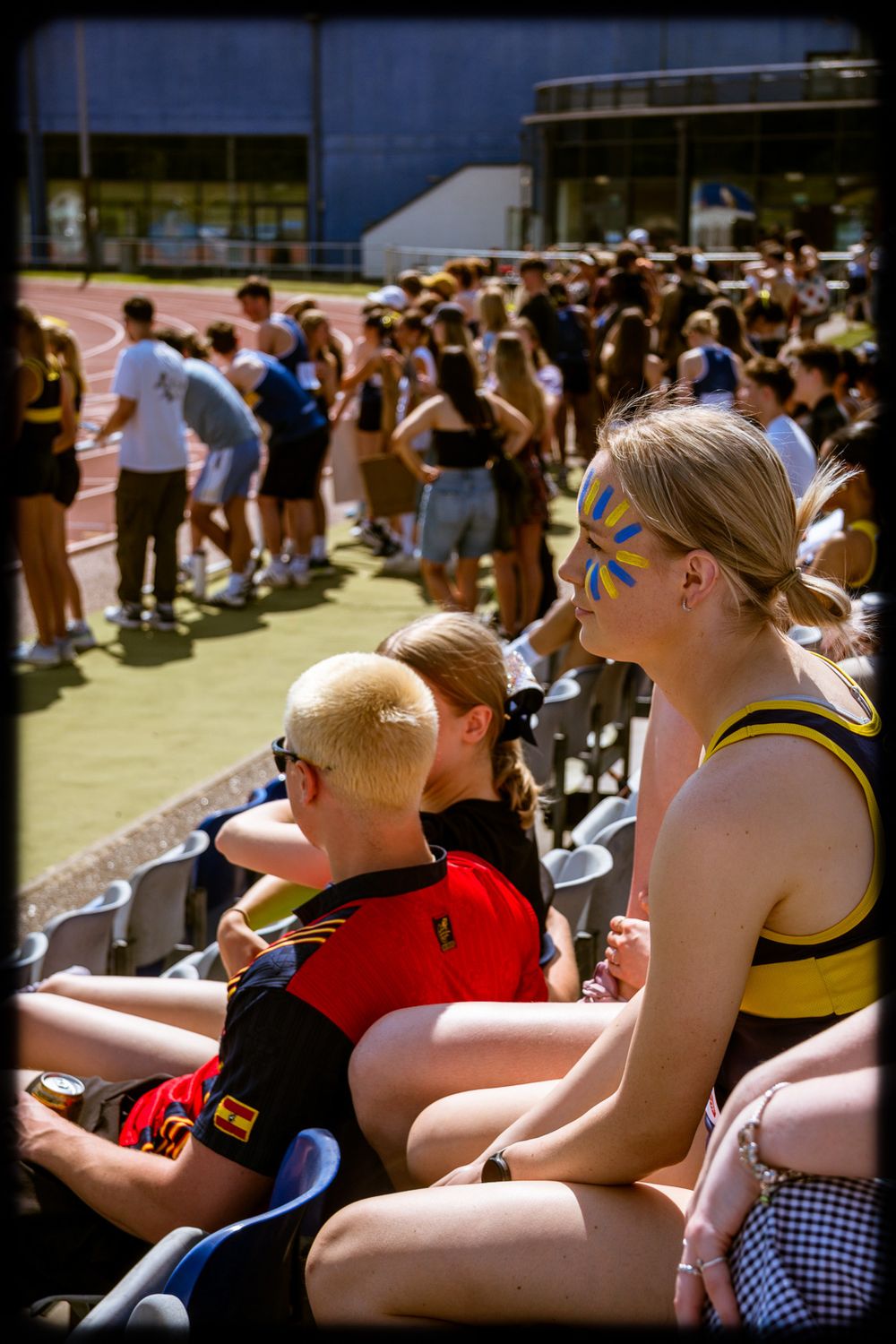 Spectators sitting in bleachers wearing sunglasses and casual attire watching an outdoor sporting event.