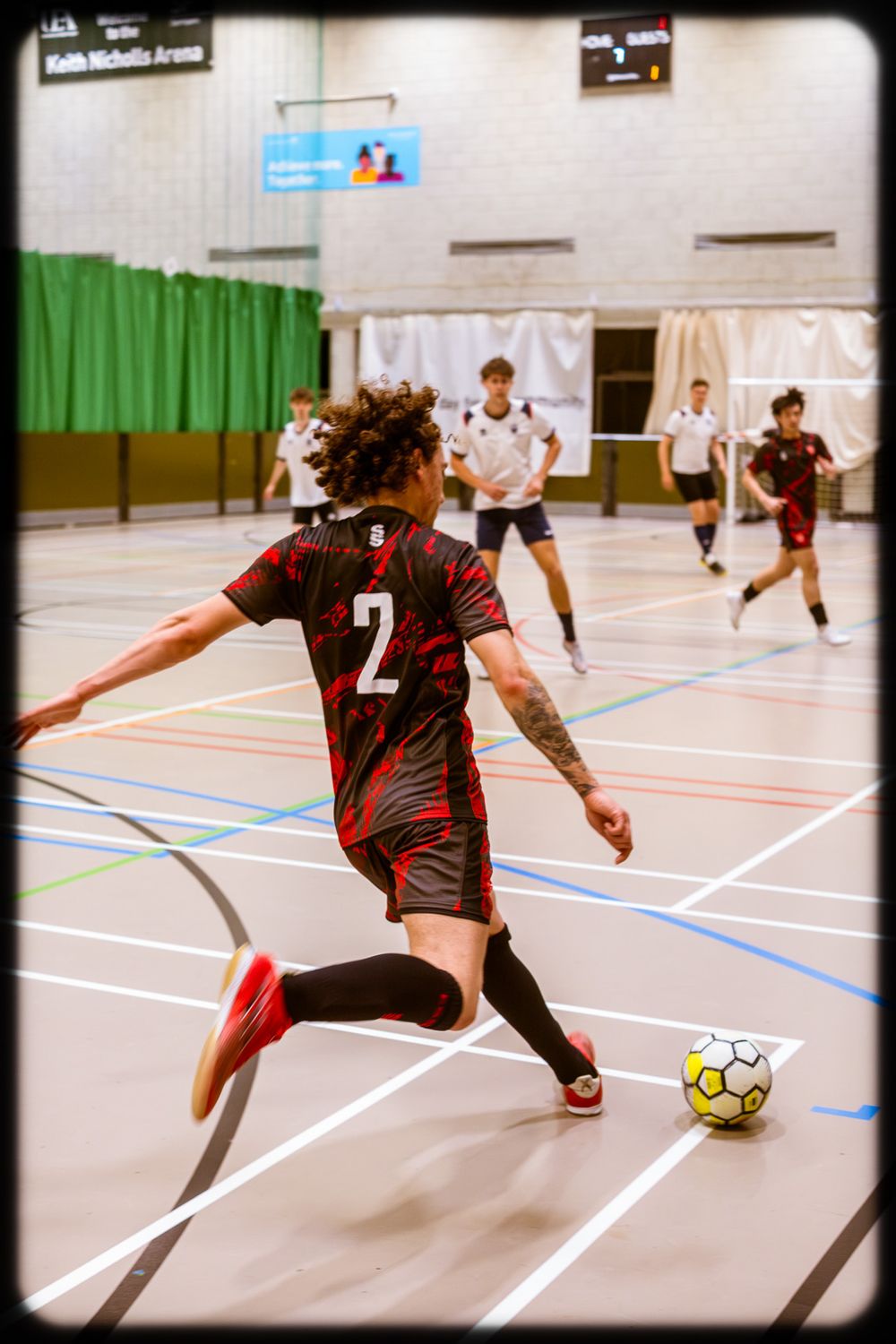 Indoor futsal player in red and black jersey number 2 dribbles ball on court during competitive match.