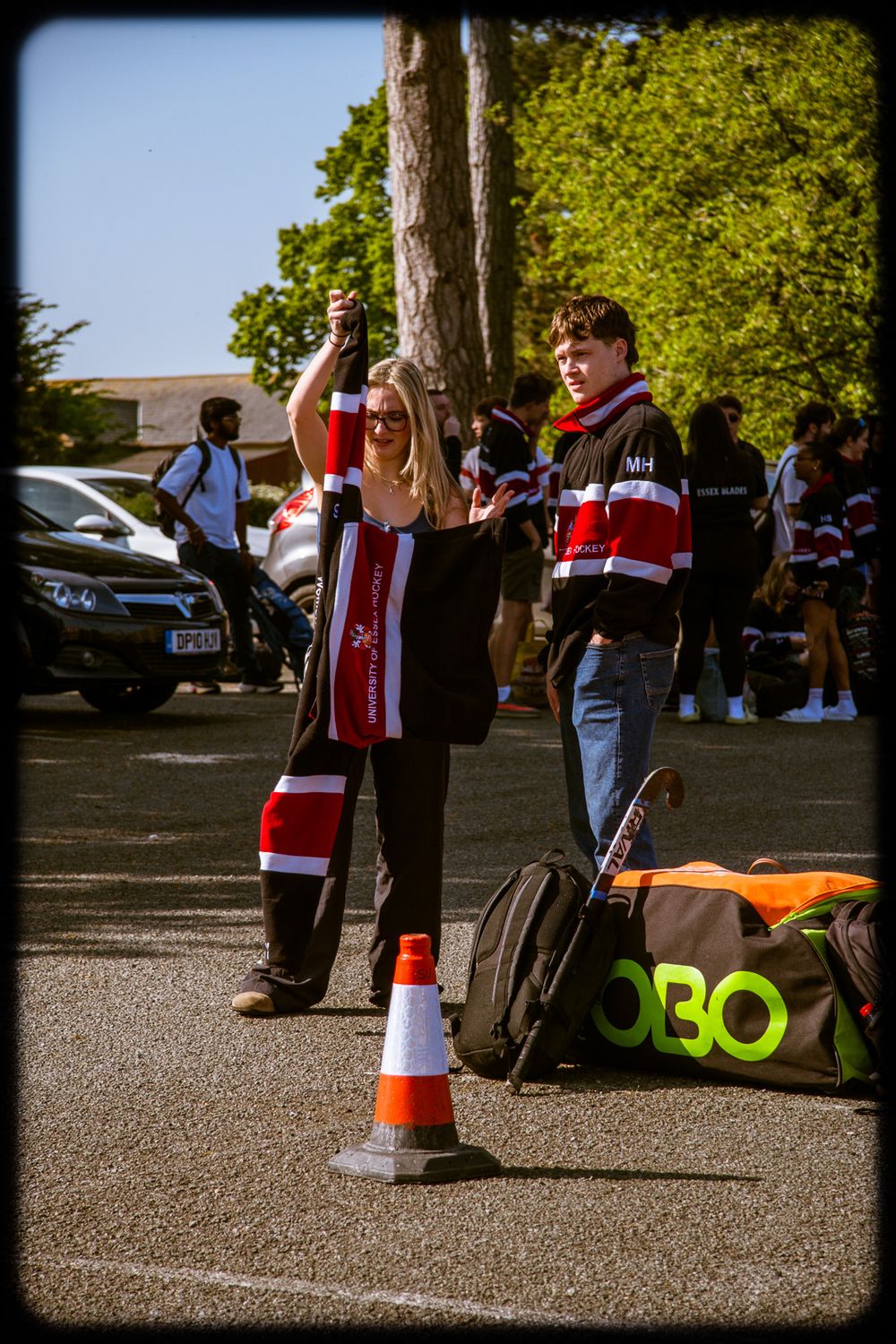 Sports fans in team scarves gather outside a stadium with traffic cones and equipment bags on a sunny day.