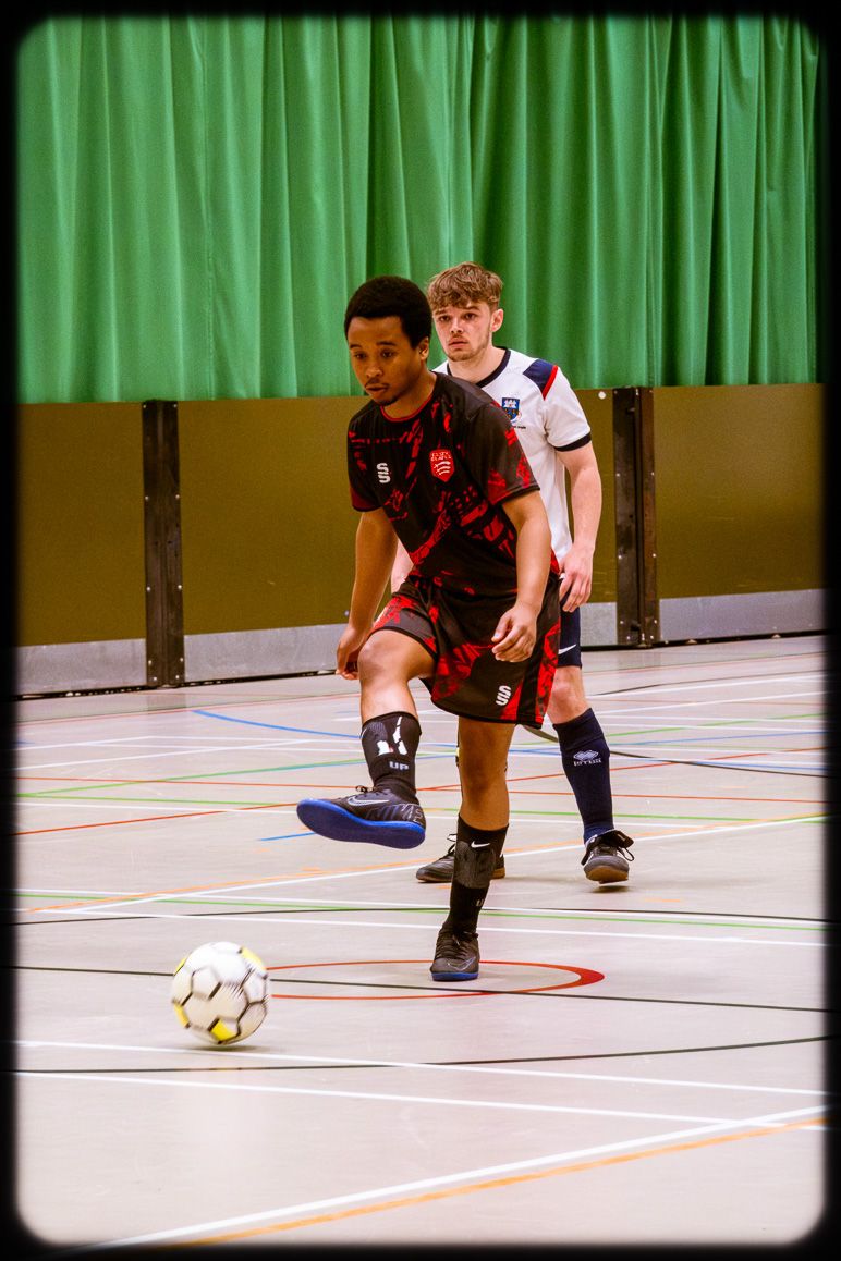 Two players compete in an indoor futsal match on a court with a green curtain backdrop and white floor markings.