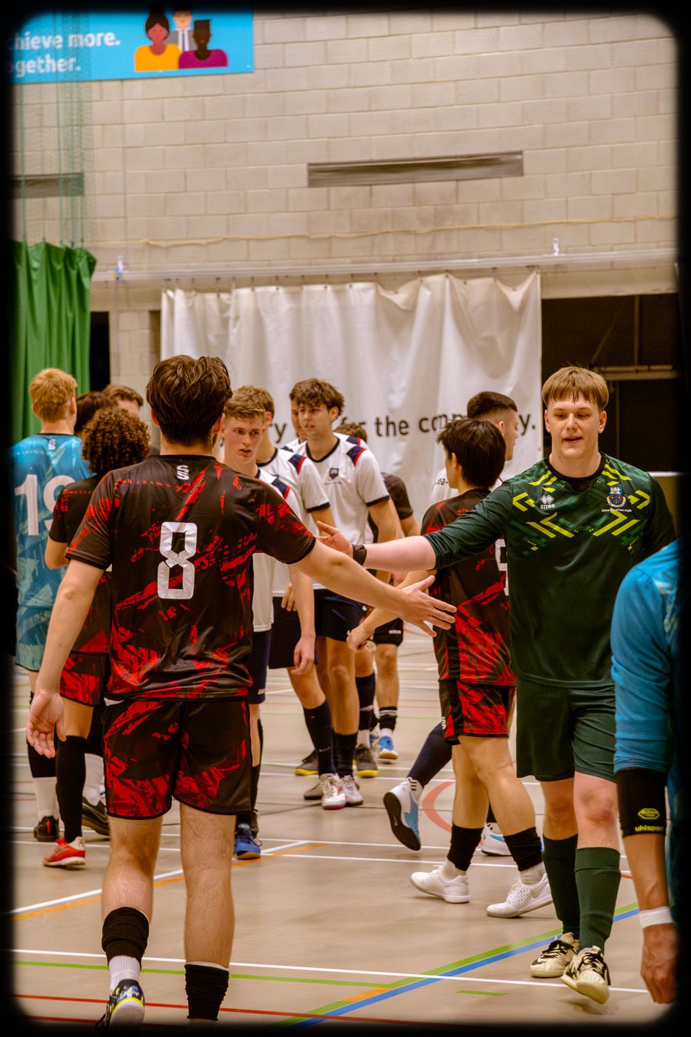 Athletes in red and black jerseys stretch and warm up together in a sports hall before a match.