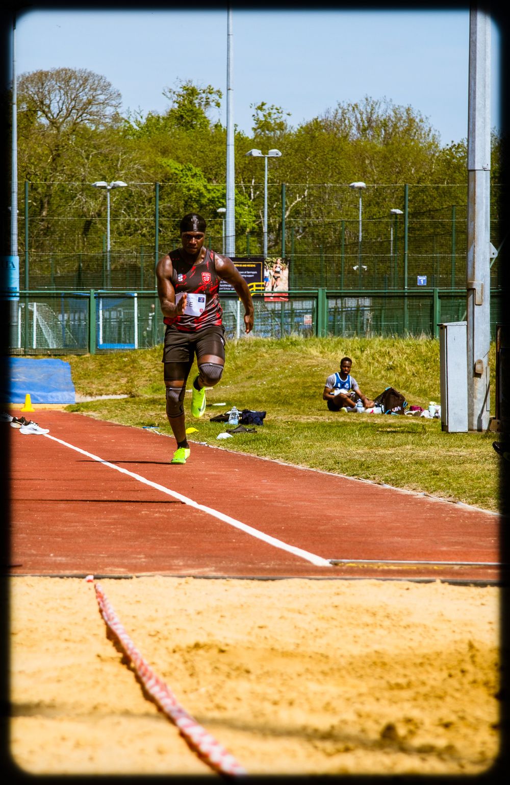 Athlete in mid-stride during long jump competition on outdoor track with sand pit landing area.