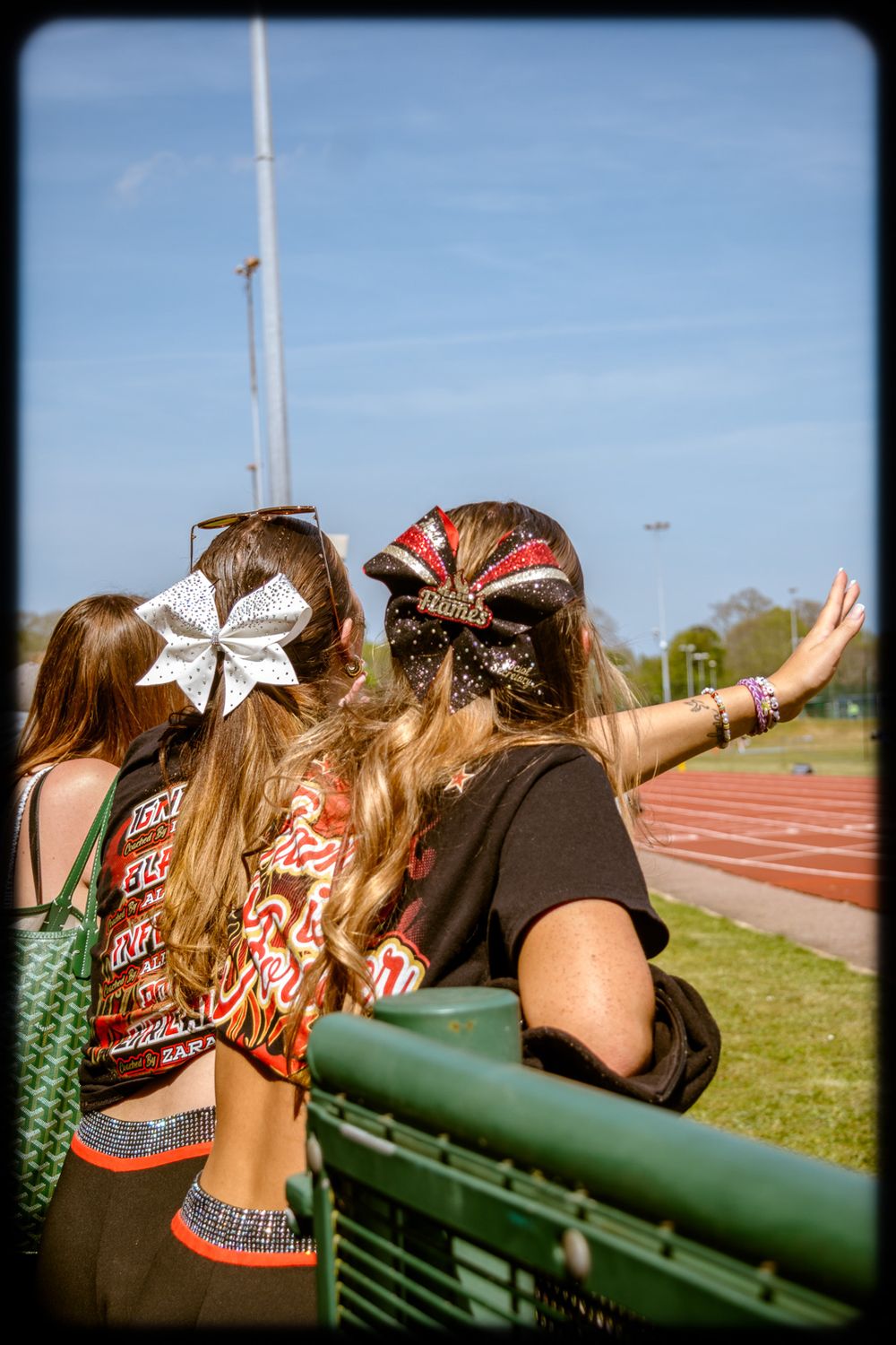 Two spectators with decorative hair bows watch from the bleachers at an outdoor track and field event.