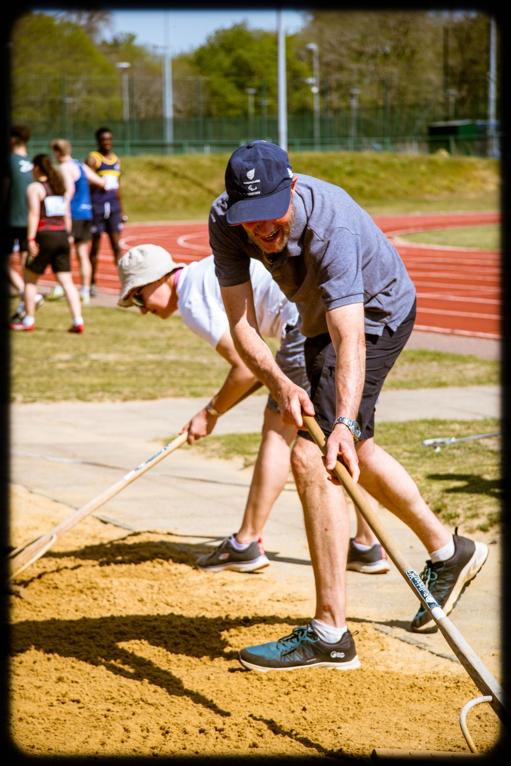 A person rakes and smooths sand in a long jump pit at an outdoor track and field facility.