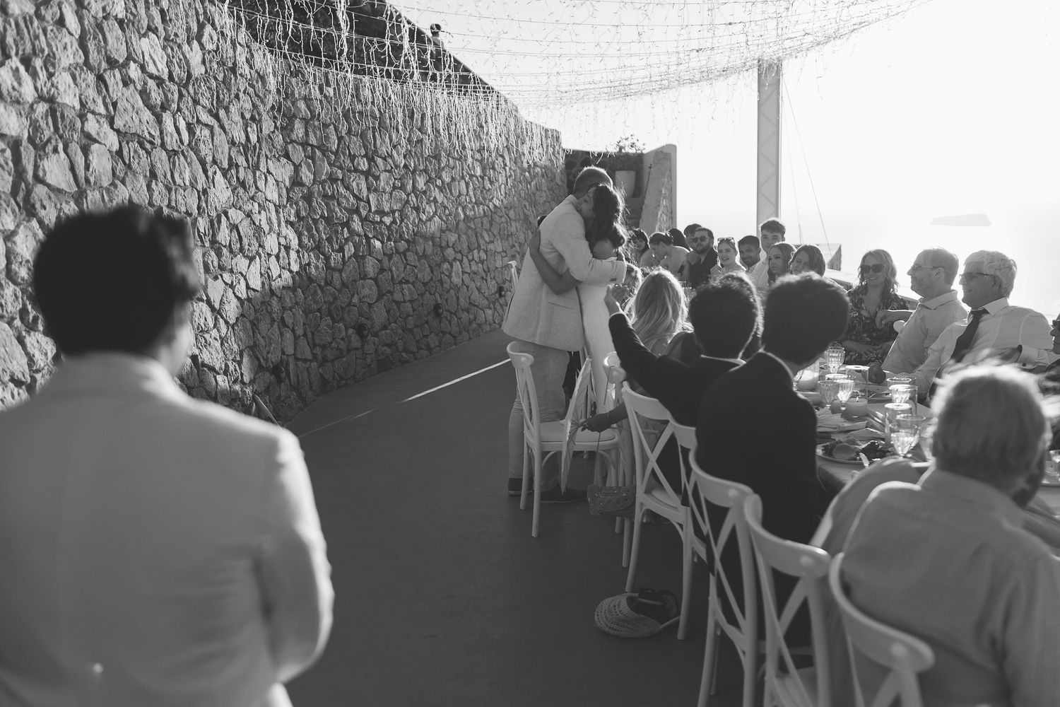 People seated in rows of white chairs attend an outdoor Santorini wedding against a stone wall backdrop.