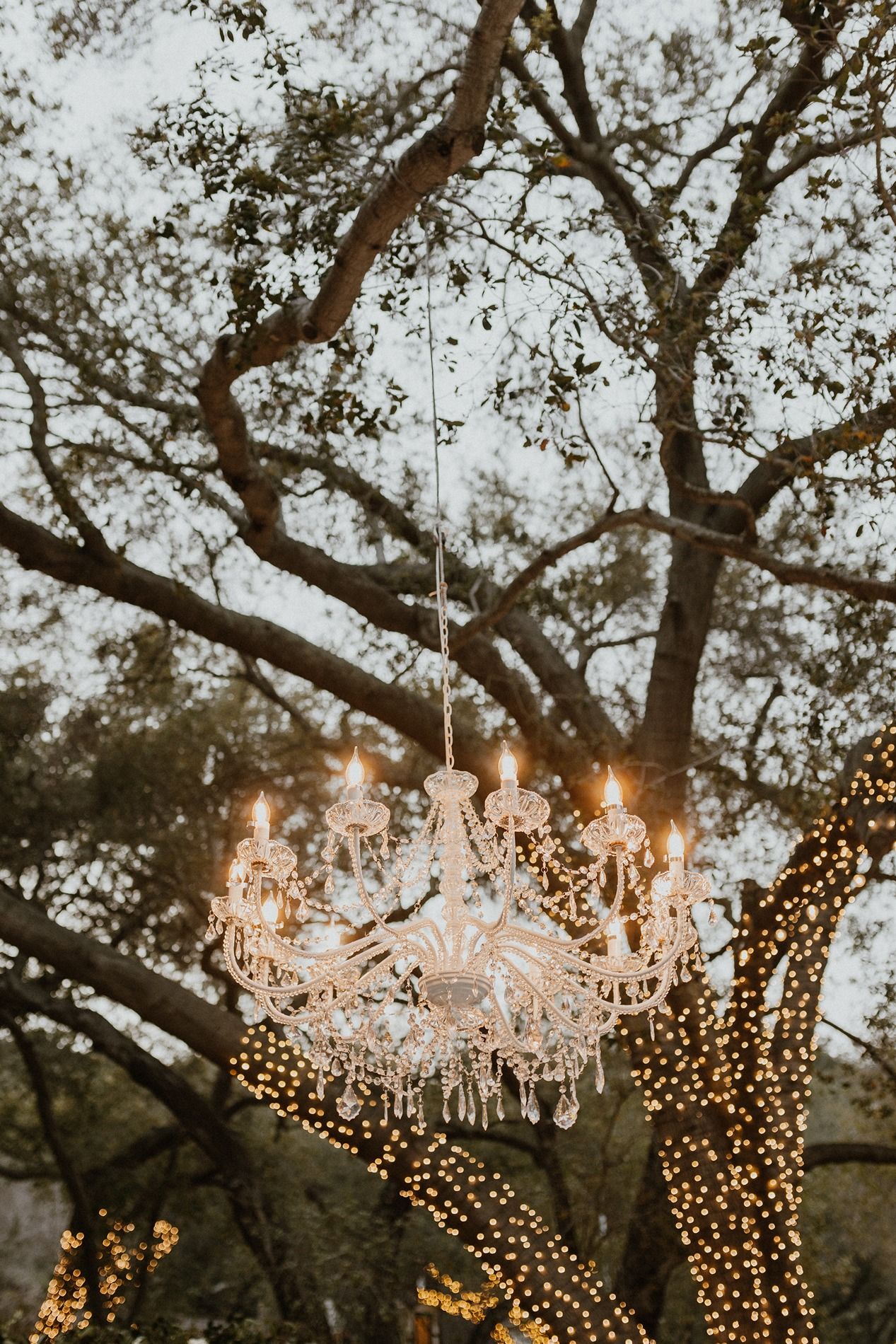 Crystal chandeliers hang from tree branches with twinkling fairy lights creating a magical outdoor setting at dusk.