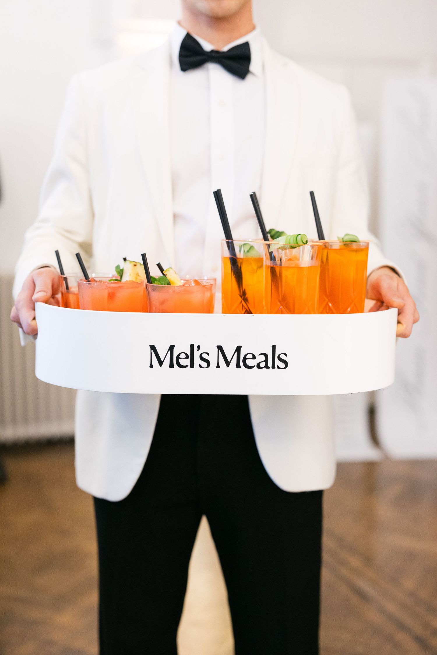 A server in formal attire carries orange cocktails on a white serving tray at an upscale event.