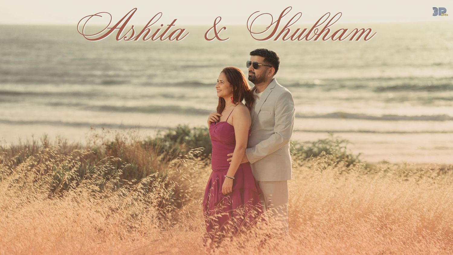 A couple stands together in tall golden grass at sunset on a beach with ocean waves in the background.