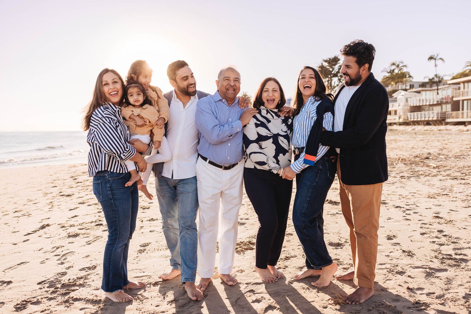 A group of people smiling together on a sunny beach with coastal homes visible in the background.