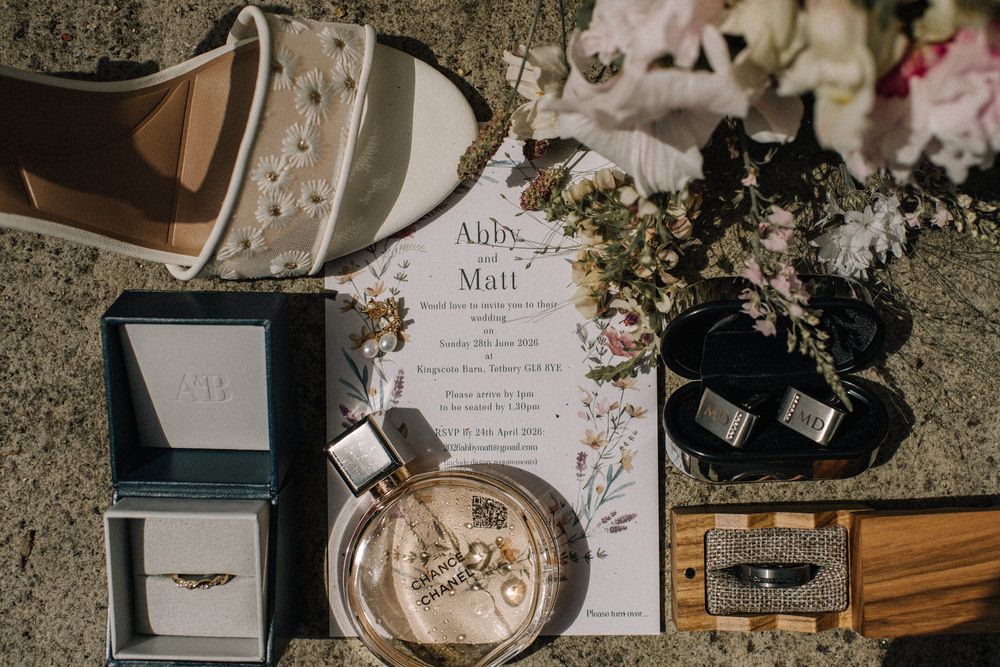 Wedding flat lay featuring invitation for Abbey and Matt, rings, perfume, bridal shoes, and floral accents on textured surface.