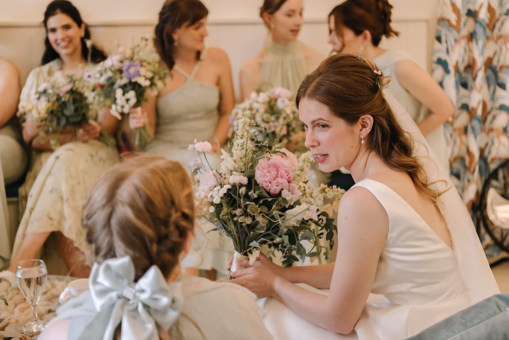 Bride in white gown smells pink floral bouquet while chatting with flower girl, bridesmaids in sage green dresses behind.