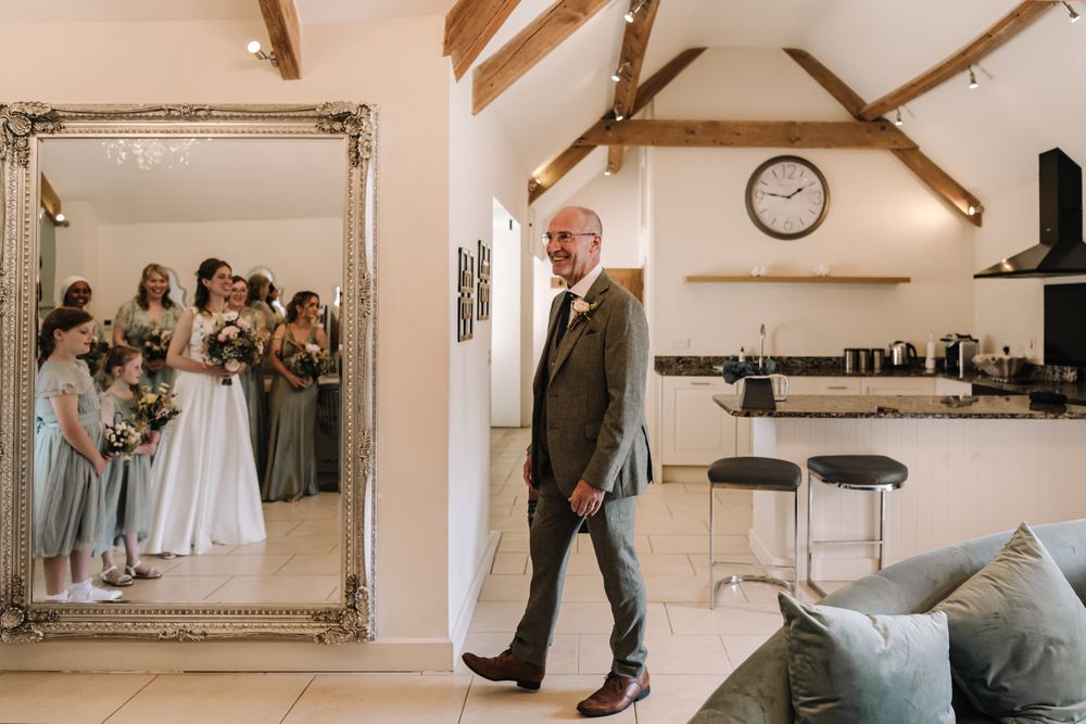 Groom in grey suit walks past large ornate mirror reflecting bridesmaids in sage dresses holding bouquets in a barn venue.