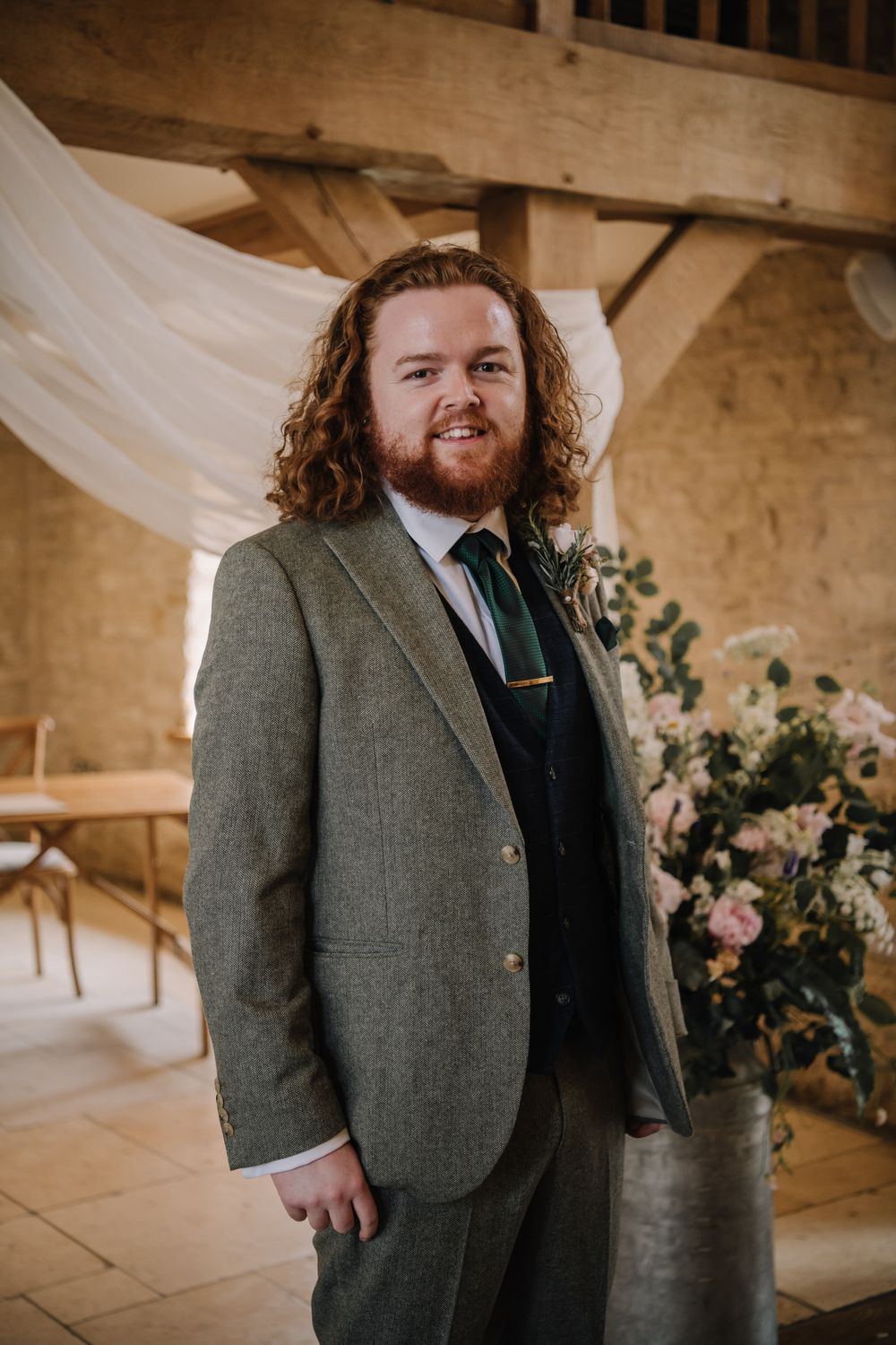 Groom in green tweed suit with dark tie and boutonniere smiling in rustic barn wedding venue.