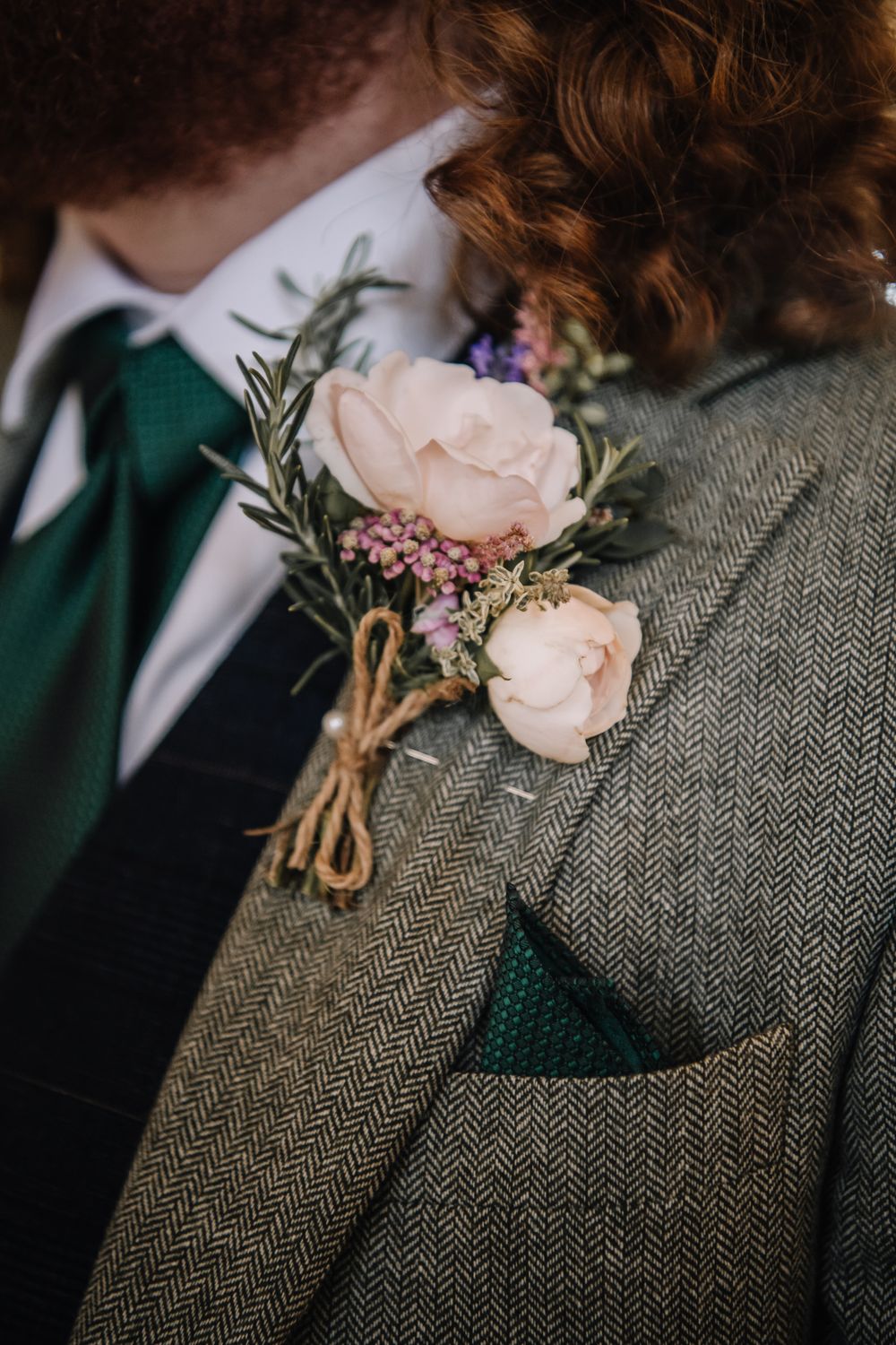 Close-up of a rustic boutonniere with blush roses and lavender on a green herringbone tweed suit jacket.