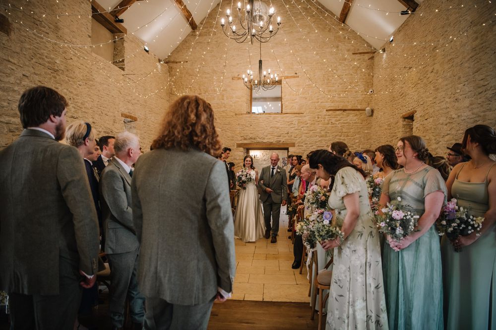 Guests watch as bride walks down the aisle in a rustic stone barn wedding venue with chandelier lighting.
