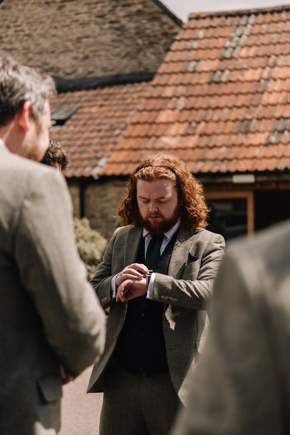 Groom with curly red hair checks his watch outdoors, wearing a green tweed suit before a wedding ceremony.